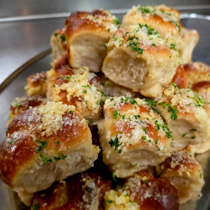 A close-up of golden-brown garlic knots, topped with parmesan cheese and fresh parsley, on a silver platter.
