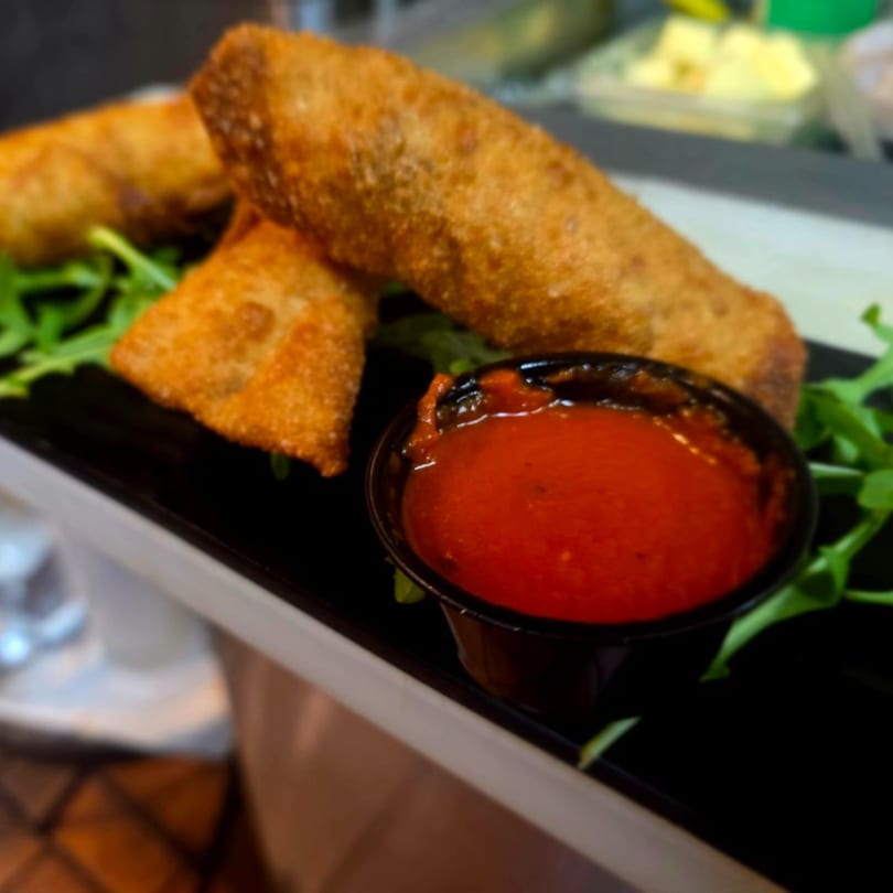 Golden-fried Italian egg rolls with a side of marinara sauce on a bed of arugula.