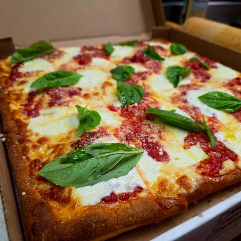 A square Buona Cucina Italiano pizza with melted mozzarella, red sauce, and fresh basil in a box.