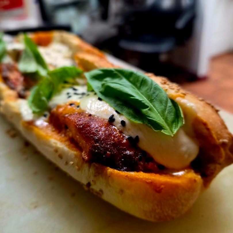 Close-up of an Italian sandwich with crispy chicken, white sauce, black sesame seeds, and basil.